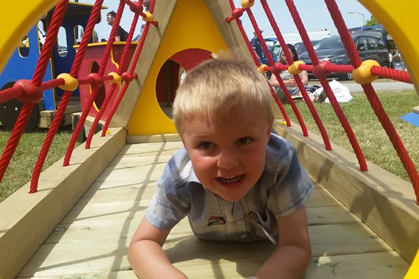 Child crawling through a wooden tunnel structure with red ropes at a playground near miniature railway cars and parked vehicles.