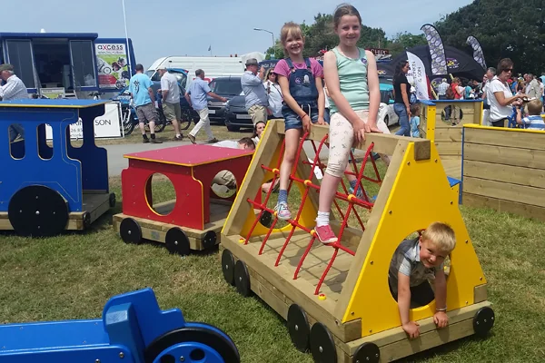 Children playing on colorful wooden trainthemed climbing structures at an outdoor event with people and vehicles in the background