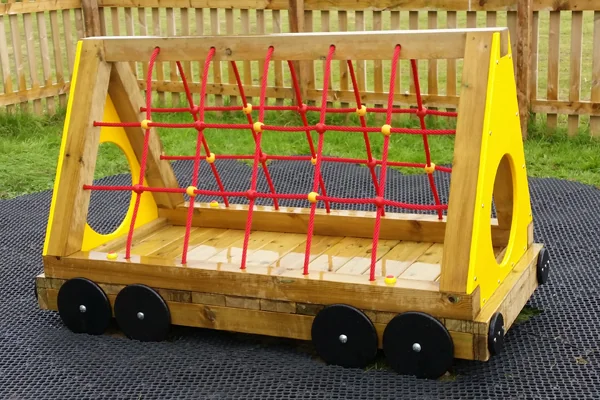 Wooden play structure shaped like a miniature train car with red climbing net and yellow side panels on black rubber matting outdoors