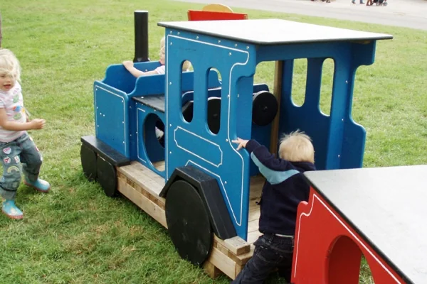 Children playing on a blue and red wooden miniature train on grass in a park setting.