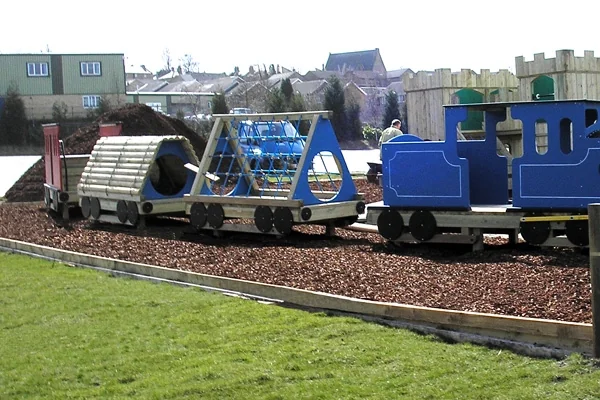 Miniature blue and wooden toy train on tracks in a playground with green grass and houses in the background