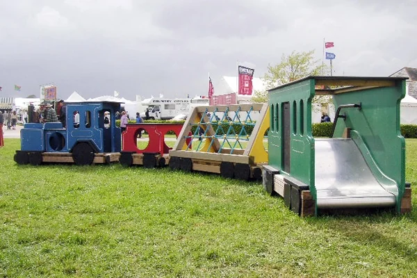Colorful outdoor wooden play train with slide and climbing frame on grass at an event