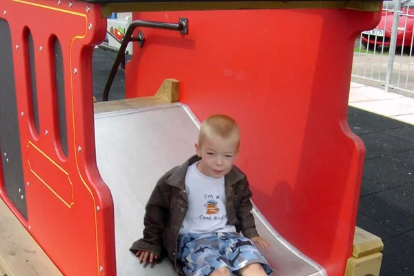 Young child sitting on a red and silver slide at a playground with a miniature railway theme.