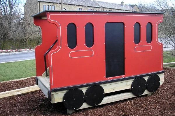Red miniature railway carriage with black wheels on wooden tracks in outdoor setting near buildings and trees