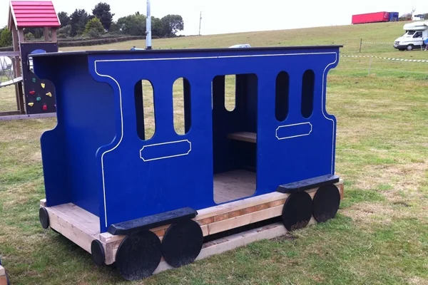 Blue wooden play train carriage on grass in an outdoor park setting with a playground in the background