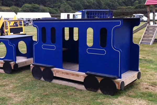 Blue wooden miniature train carriage with black wheels on grass in an outdoor play area with playground equipment in background