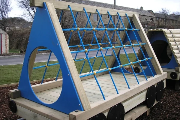 Wooden climbing frame with blue rope netting on a miniature railway platform outdoors near a road and buildings.