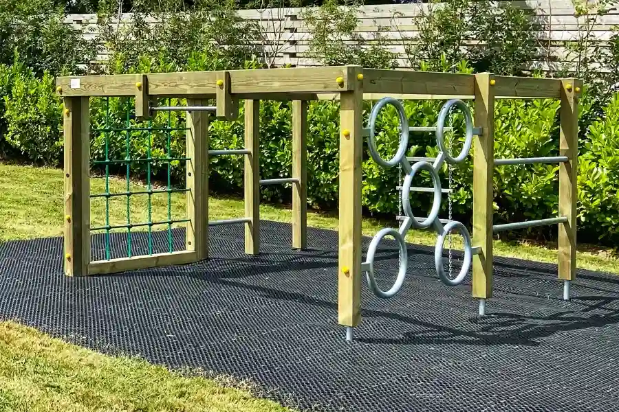 The Delabole Activity Centre timber climbing frame with a green rope net and steel monkey rings, set on black rubber matting in a landscaped outdoor playground with hedges.