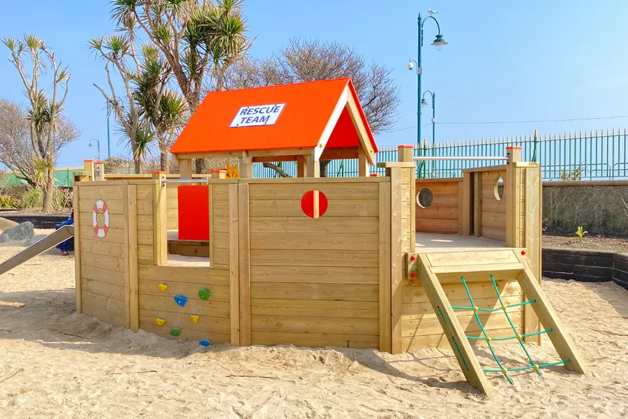 A wooden lifeboat play structure with a red roof and "RESCUE TEAM" sign, set in a sandy beach playground.