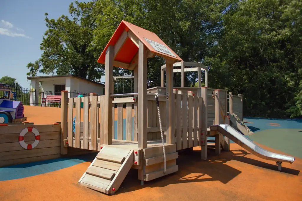 Wooden children's play structure with orange roof, rope ladder, small slide and colourful safety surfacing