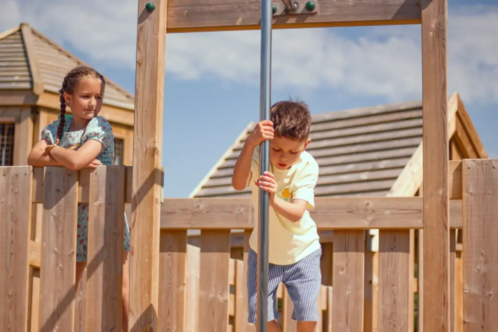 Young boy holding a vertical metal pole on a wooden playframe while a girl watches from the platform.