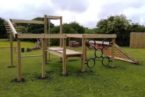 The Torpoint Activity Centre, a timber climbing frame with monkey rings, a red rope net, a climbing wall, and overhead ladders, set on rubber mulch in a grassy outdoor playground.